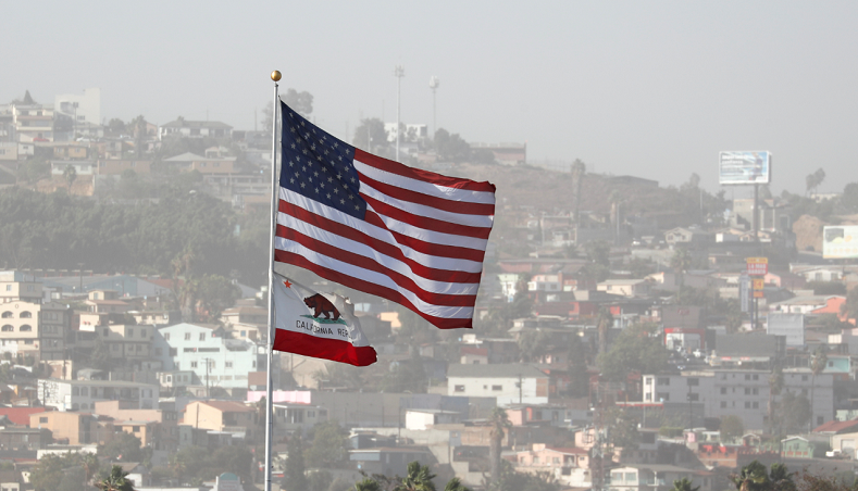 U.S. and California state flags at the Tijuana-San Diego border. The approximately 10,000 migrants from four Central American caravans are expected to arrive here in the coming days. About 70 LGBTQ migrants arrived in Tijuana Sunday.