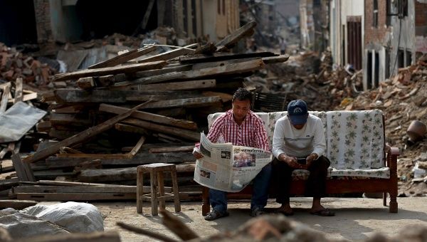 A man reads a newspaper as his friend looks at his mobile phone, as they sit next to collapsed houses on the outskirts of Kathmandu, Nepal, May 15, 2015.