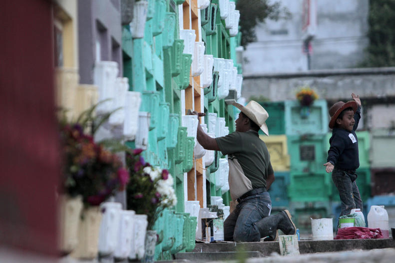 Solola, Guatemala. A man and his son repaint an above ground grave. People wait for the first rays of the sun to illuminate the colorful tombstones, and gather in cemeteries with fresh flowers and food.  