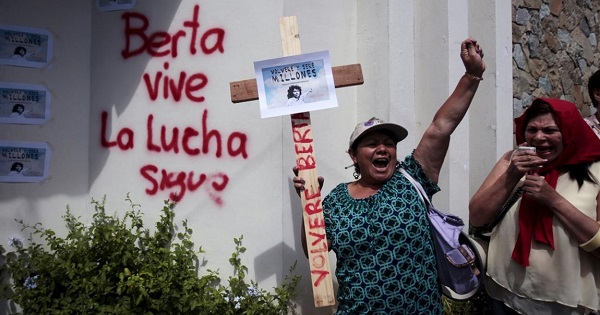 A woman holds a cross with a sign featuring Berta Caceres that says, 