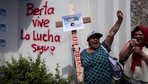 A woman holds a cross with a sign featuring Berta Caceres that says, 