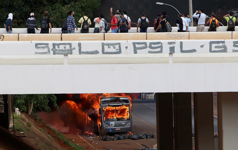 People observe a burning bus from a nearby bridge.