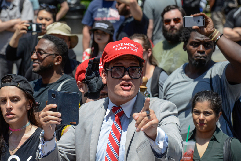A man wearing a Make America Great Again cap gestures while people participate in an event called 