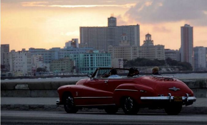 Sunsets over Havana's Malecon as tourists ride in a convertible car.