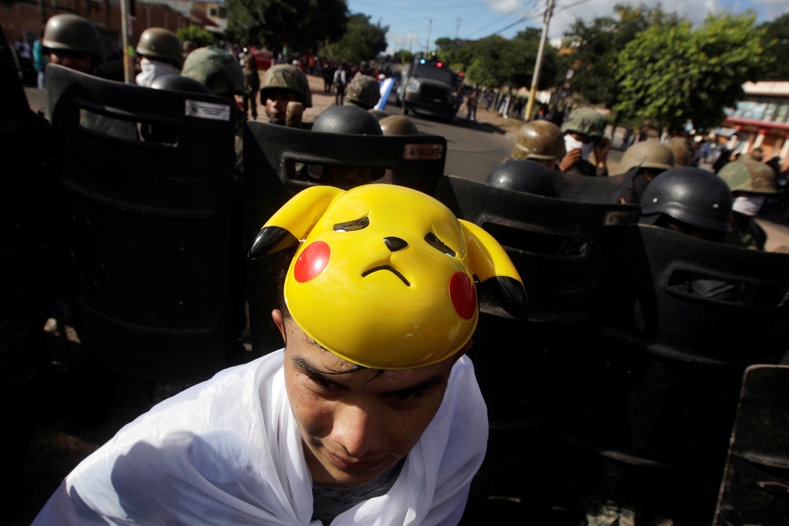A man wearing a Pikachu mask turns his back to heavily armed riot police.