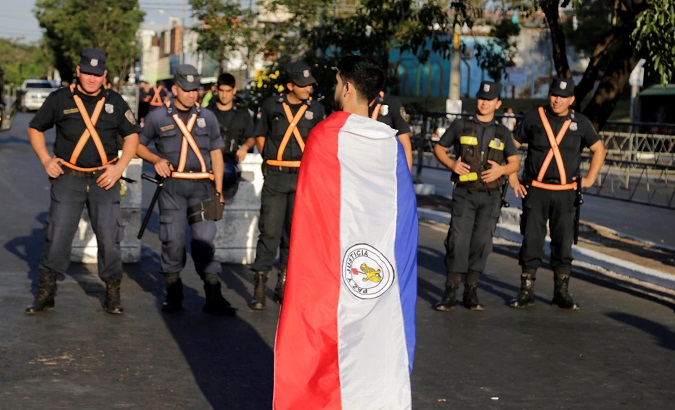 Paraguayan protester stands wrapped in national flag facing state security forces.