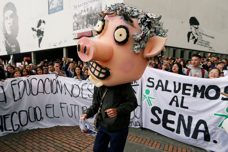 A protester with a mask depicting President Duque. In the background, people hold signs reading: 