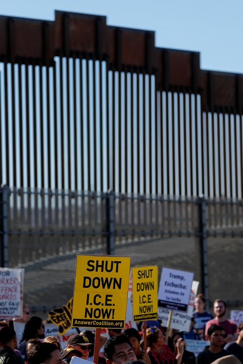 Along the border wall, proesters hold up signs reading 