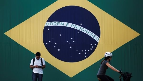 Man checks his mobile phone as a woman riding a bike passes next to a Brazilian flag in Sao Paulo, Brazil June 28, 2018.